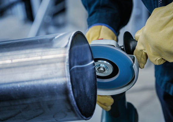 Person wearing safety equipment grinds a large metal pipe with an angle grinder.
