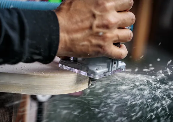 Person shaping wood with a handheld power router, creating dust and shavings.