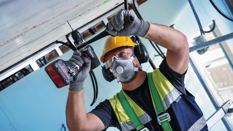 Person wearing safety equipment drills into a ceiling at a construction site.