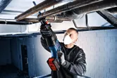 A person wearing safety equipment uses a cordless reciprocating saw to cut overhead pipes.