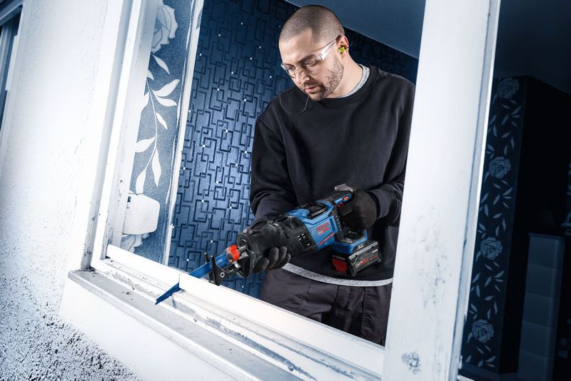 A person wearing safety equipment removes a window frame with a cordless reciprocating saw.