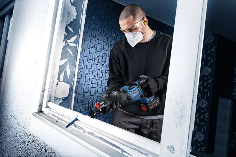 A person wearing safety equipment uses a cordless reciprocating saw to cut a window frame.