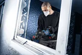 A person wearing safety equipment uses a cordless reciprocating saw to cut a window frame.