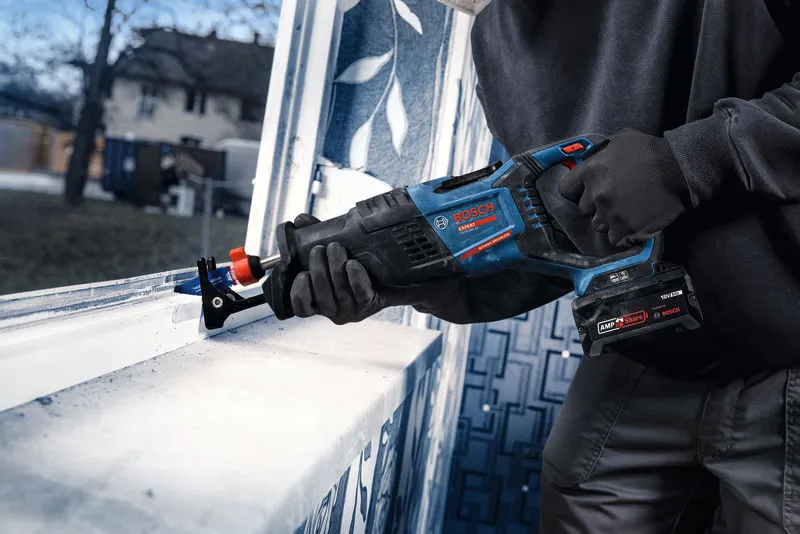 A person wearing safety equipment cuts a window frame with a cordless reciprocating saw.