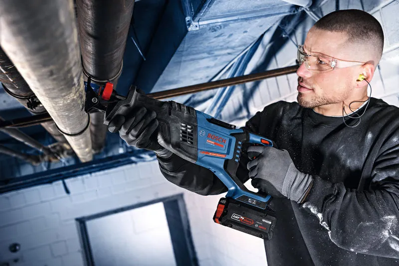 A person wearing safety equipment uses a cordless reciprocating saw to cut overhead pipes.