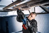 A person wearing safety equipment uses a cordless reciprocating saw to cut overhead pipes.
