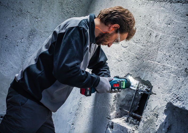 A person wearing safety equipment uses a cordless multi-cutter on a concrete wall.
