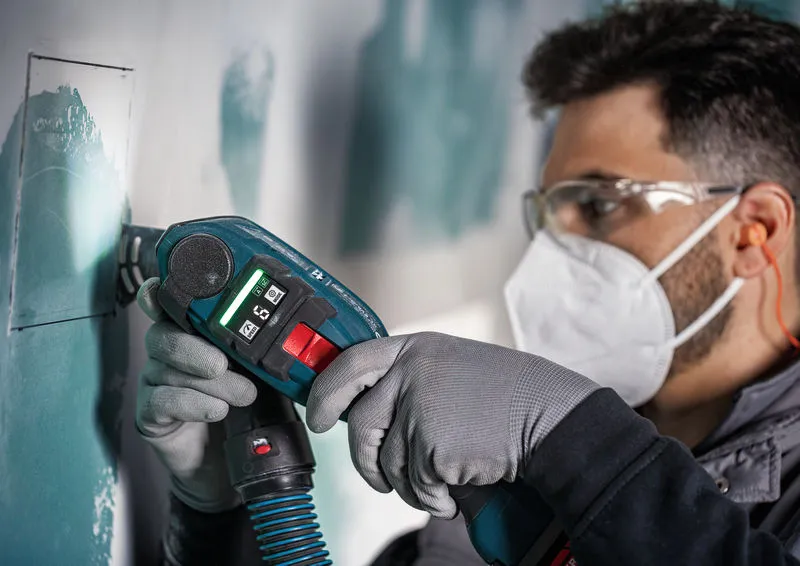 A person wearing safety equipment uses a cordless multi-cutter to cut drywall.