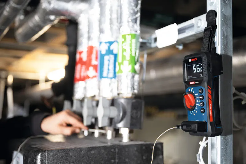 A person inspects pipes while a digital multimeter measures temperature nearby.