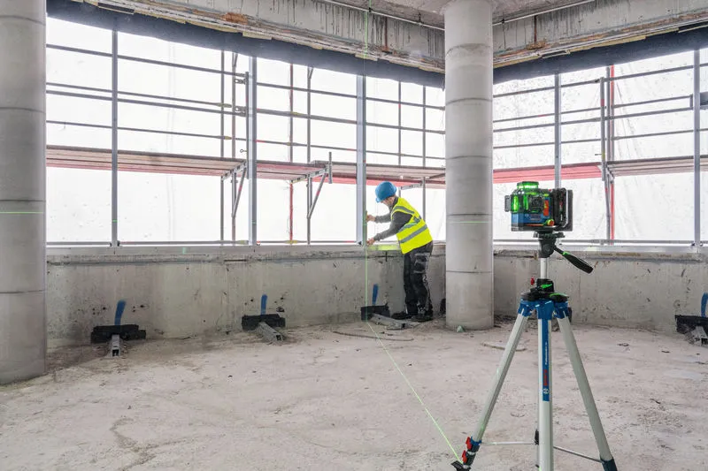 A person wearing safety equipment aligns window frames using a laser leveling tool on a tripod.