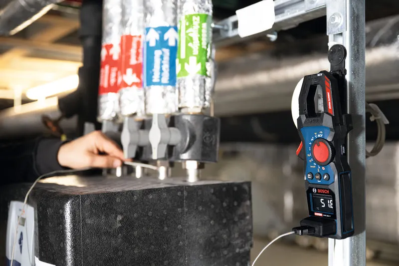 A person works near insulated pipes using a clamp meter in an industrial setting.
