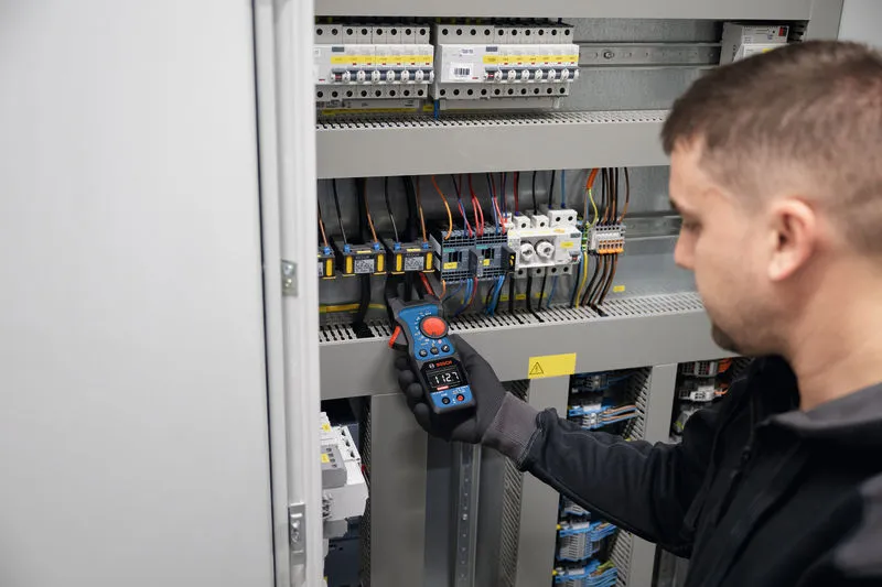 A person wearing safety equipment measures voltage in an electrical panel with a clamp meter.