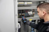A person wearing safety equipment measures voltage in an electrical panel with a clamp meter.