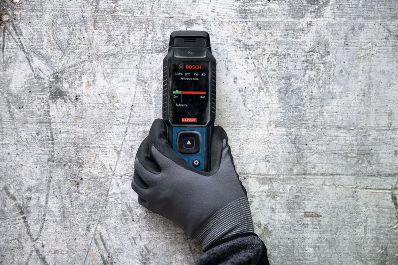 A person wearing safety equipment uses a material moisture meter on a concrete wall.