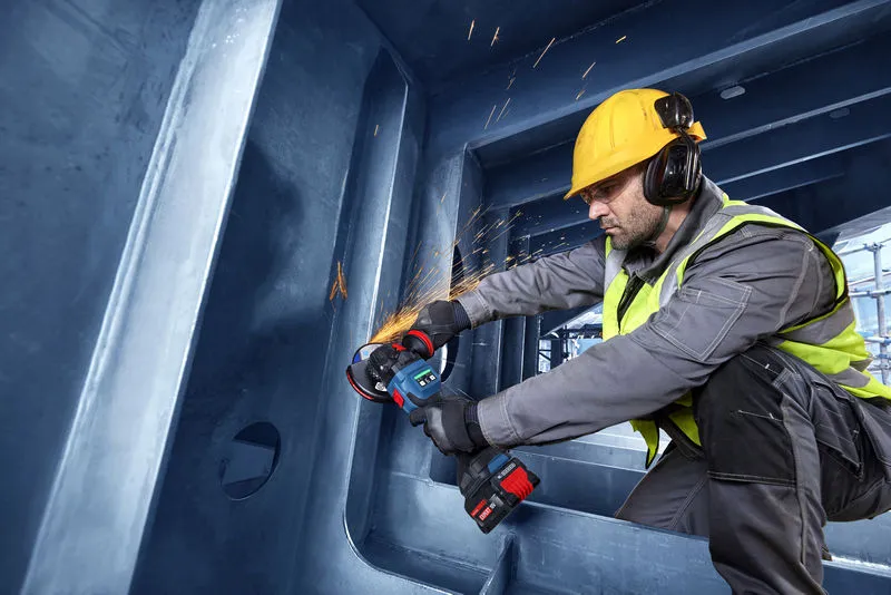 A person wearing safety equipment uses an angle grinder to cut metal inside a large structure.