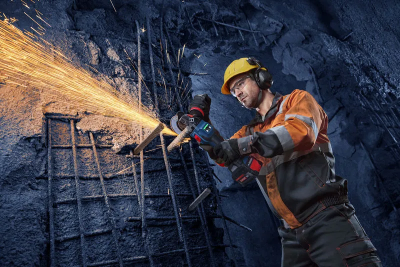 A person wearing safety equipment uses an angle grinder to cut metal rods, producing sparks.