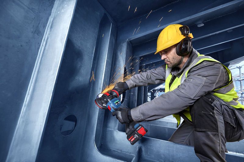 A person wearing safety equipment uses a cordless angle grinder on a steel structure.