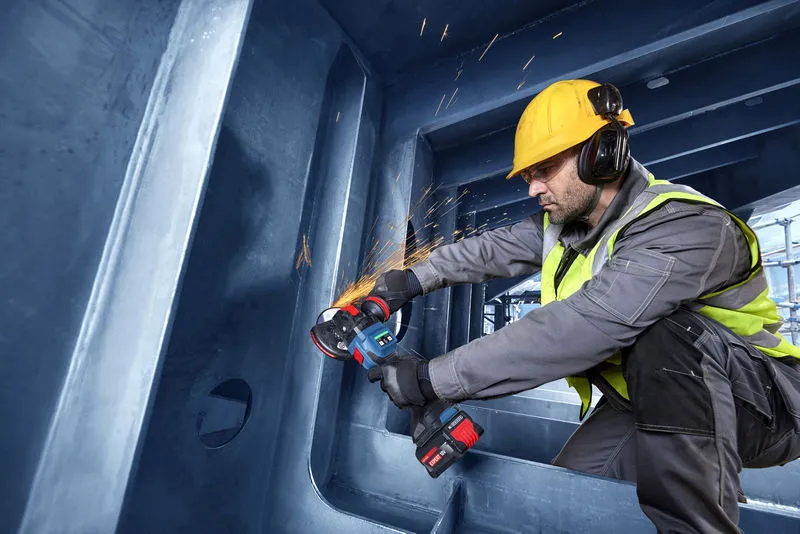 A person wearing safety equipment uses a cordless angle grinder on a steel structure.