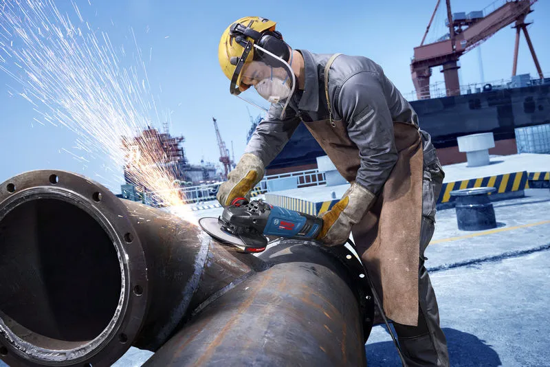A worker wearing safety equipment grinds a large metal pipe, sending sparks flying.