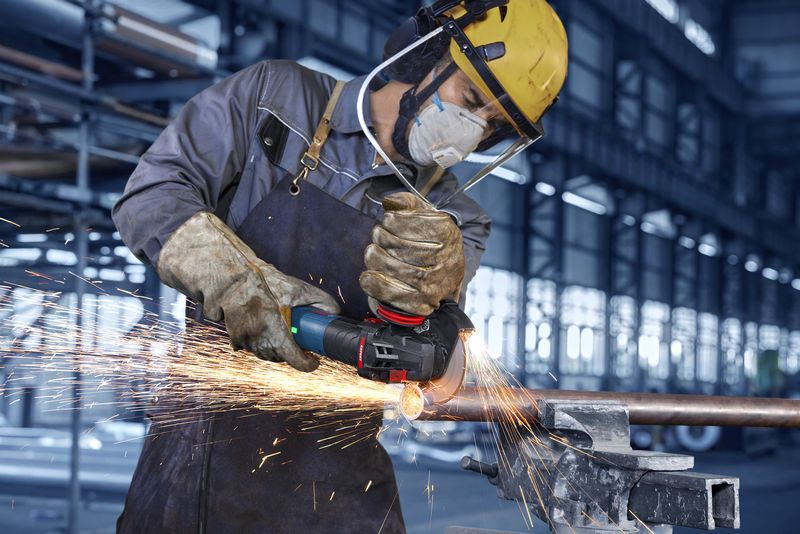 A person wearing safety equipment grinds a metal pipe, creating bright sparks.