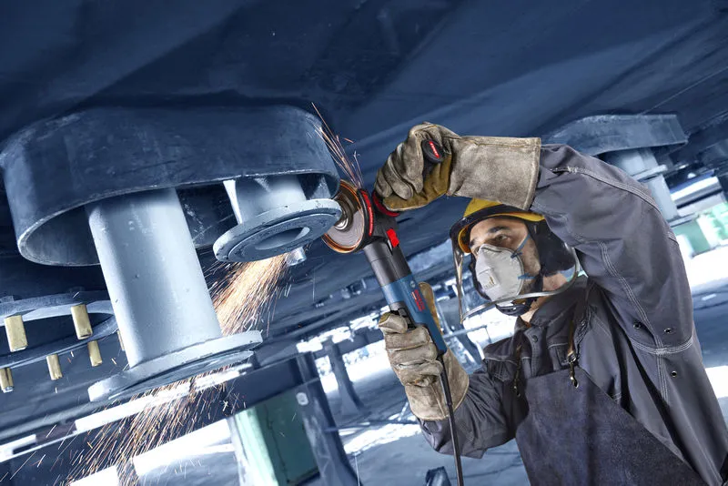 A person wearing safety equipment grinds metal overhead with an angle grinder, creating sparks.