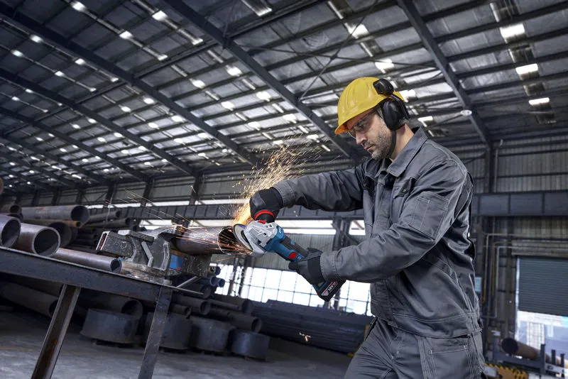 A person wearing safety equipment uses an angle grinder on a metal pipe, creating sparks.