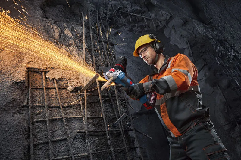 A person wearing safety equipment uses an angle grinder to cut rebar, with sparks flying.