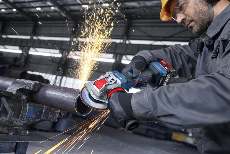 A person wearing safety equipment grinds a metal pipe with a cordless angle grinder.