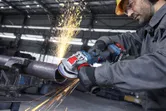 A person wearing safety equipment grinds a metal pipe with a cordless angle grinder.