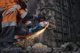 A person wearing safety equipment grinds concrete with a cordless angle grinder.