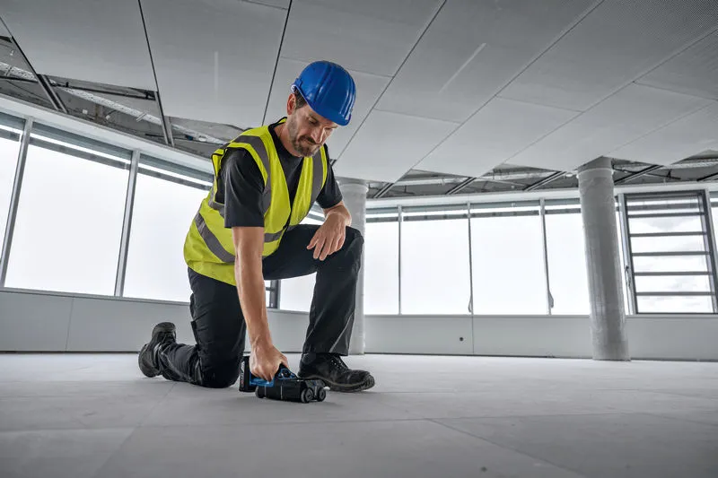 A person wearing safety equipment uses a detector to scan the floor of a large room.