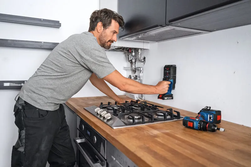A person uses a detector on a kitchen wall above a countertop.
