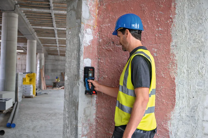 A person wearing safety equipment uses a wall detector on a concrete column.