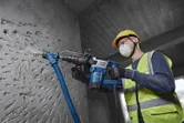 A worker wearing safety equipment drills into a concrete wall using a power tool with dust extraction.