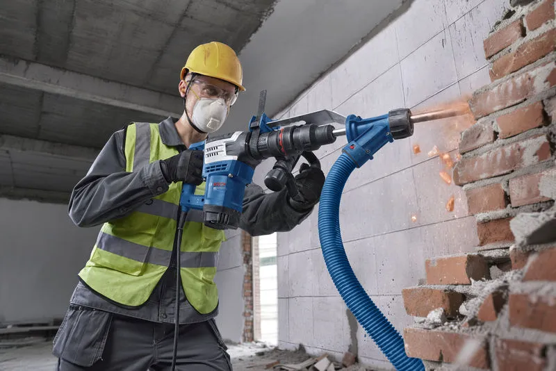A person wearing safety equipment uses a rotary hammer drill on a brick wall.