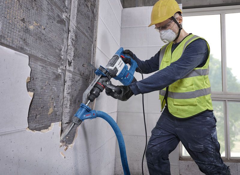 A person wearing safety equipment removes wall tiles with a power tool connected to a vacuum hose.