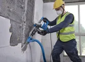 A person wearing safety equipment removes wall tiles with a power tool connected to a vacuum hose.