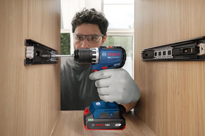 A person wearing safety equipment uses a cordless drill driver to assemble a wooden cabinet.