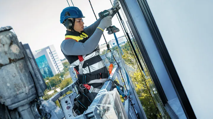 A person wearing safety equipment uses a power drill on a building facade.