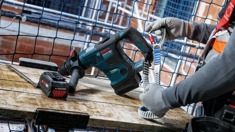 Person wearing safety equipment attaches a lanyard to a cordless demolition tool on a workbench.