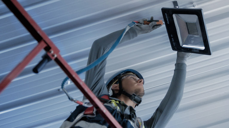 Person wearing safety equipment installs a floodlight on a metal ceiling.