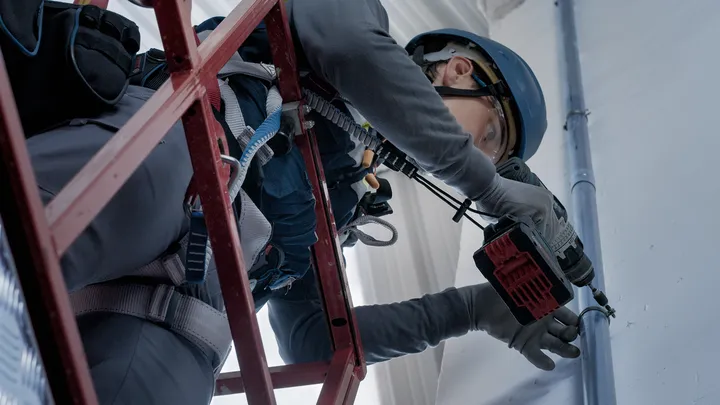 Person wearing safety equipment uses a power tool to drill into a wall from a scaffold.
