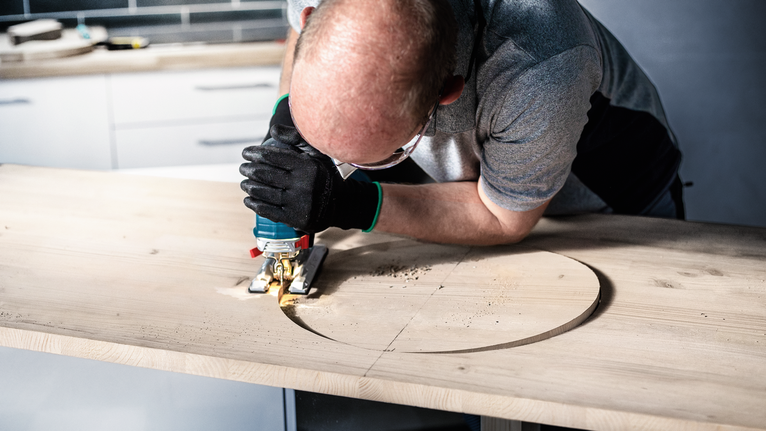 Person wearing safety equipment uses a jigsaw to cut a circle in a wooden board.