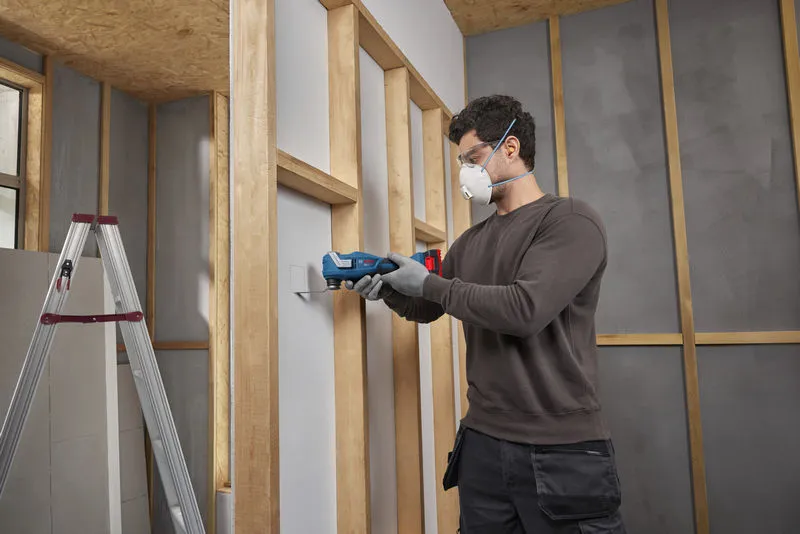 A person wearing safety equipment uses a multifunction tool to cut drywall.
