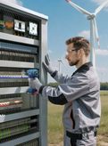 A person wearing safety equipment uses a cordless tool on an electrical cabinet outdoors.