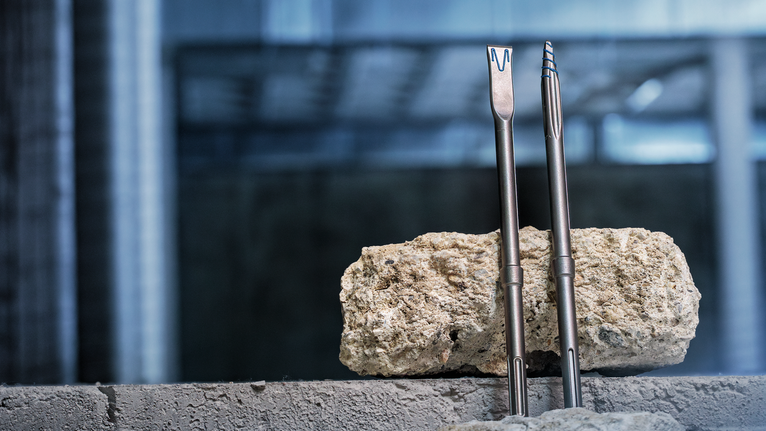 Two chisel bits stand upright against a rough stone block on a concrete surface.