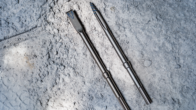 Two metal chisel drill bits lie on rough concrete.