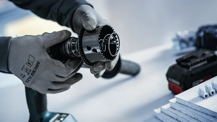 Person wearing safety equipment attaches a hole saw to a drill near stacked boards.