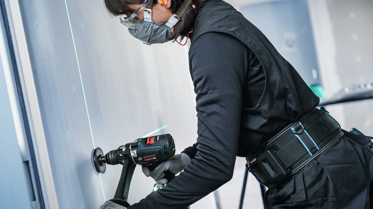 Person wearing safety equipment drills into a wall guided by a green laser line.