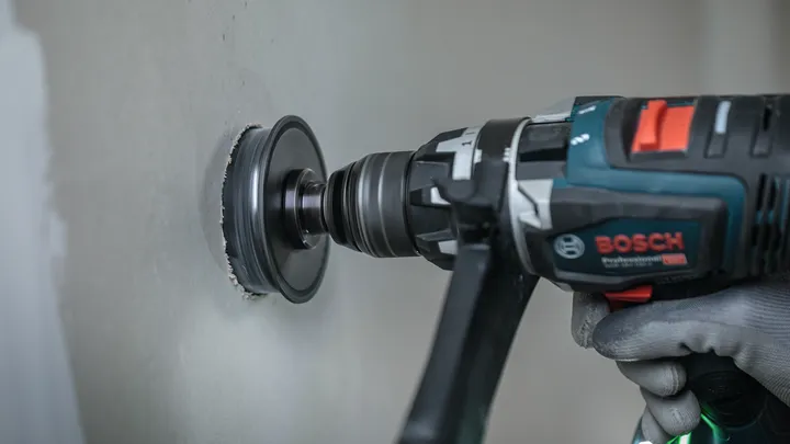Person wearing safety equipment drills a circular hole into a drywall panel.